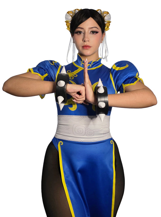 Photo of a model wearing the Chun-Li classic blue qipao cosplay costume from Street Fighter, with gold hair buns (ox horns), white waist belt, and black spiked wrist cuffs. The model is posing with hands clasped.