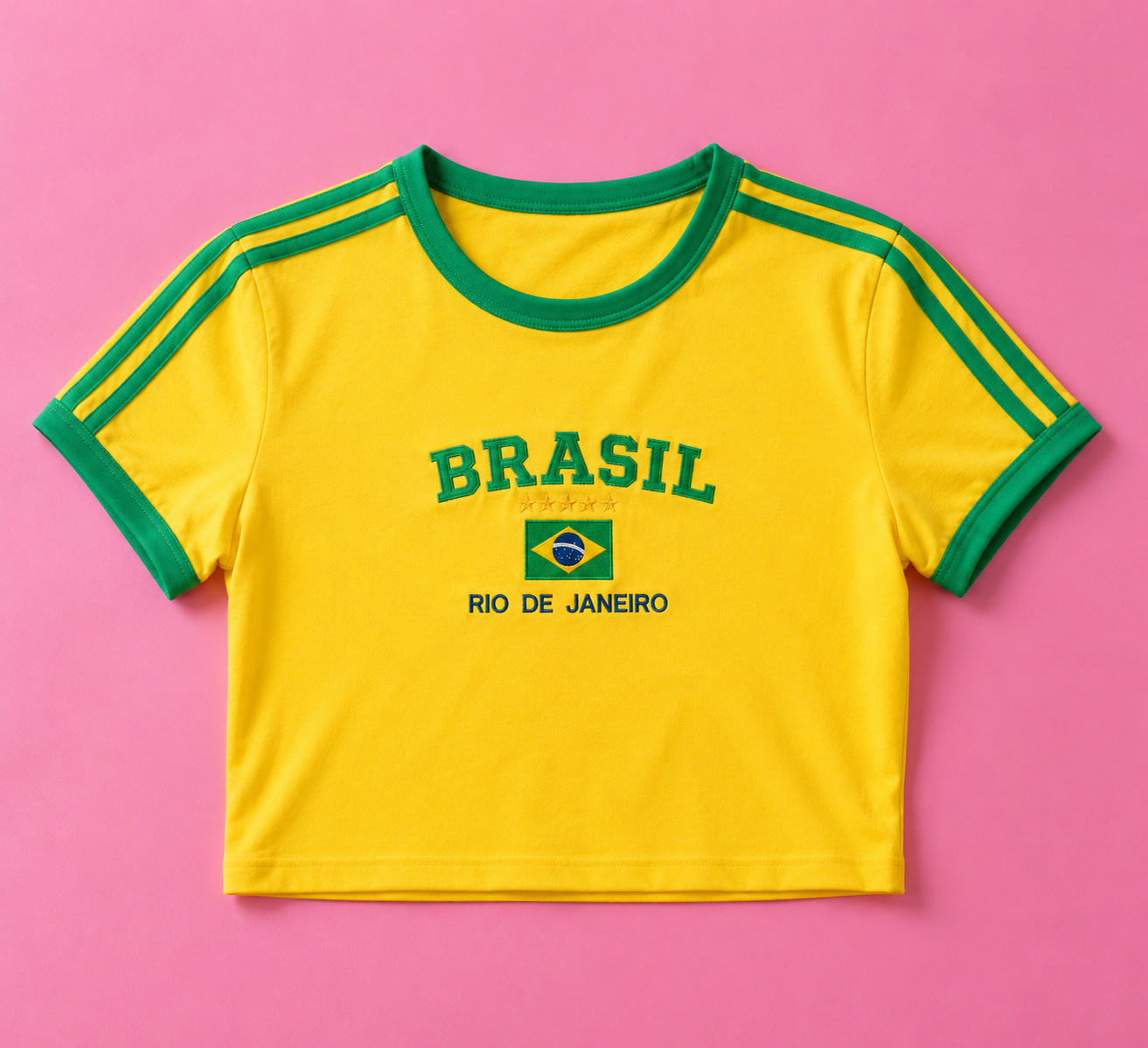 A yellow ringer crop top t-shirt with green trim on the collar and sleeves, featuring 'BRASIL RIO DE JANEIRO' and a small Brazilian flag printed on the chest, laid flat on a pink background.