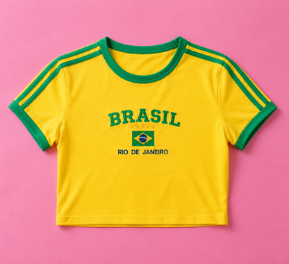 A yellow ringer crop top t-shirt with green trim on the collar and sleeves, featuring 'BRASIL RIO DE JANEIRO' and a small Brazilian flag printed on the chest, laid flat on a pink background.