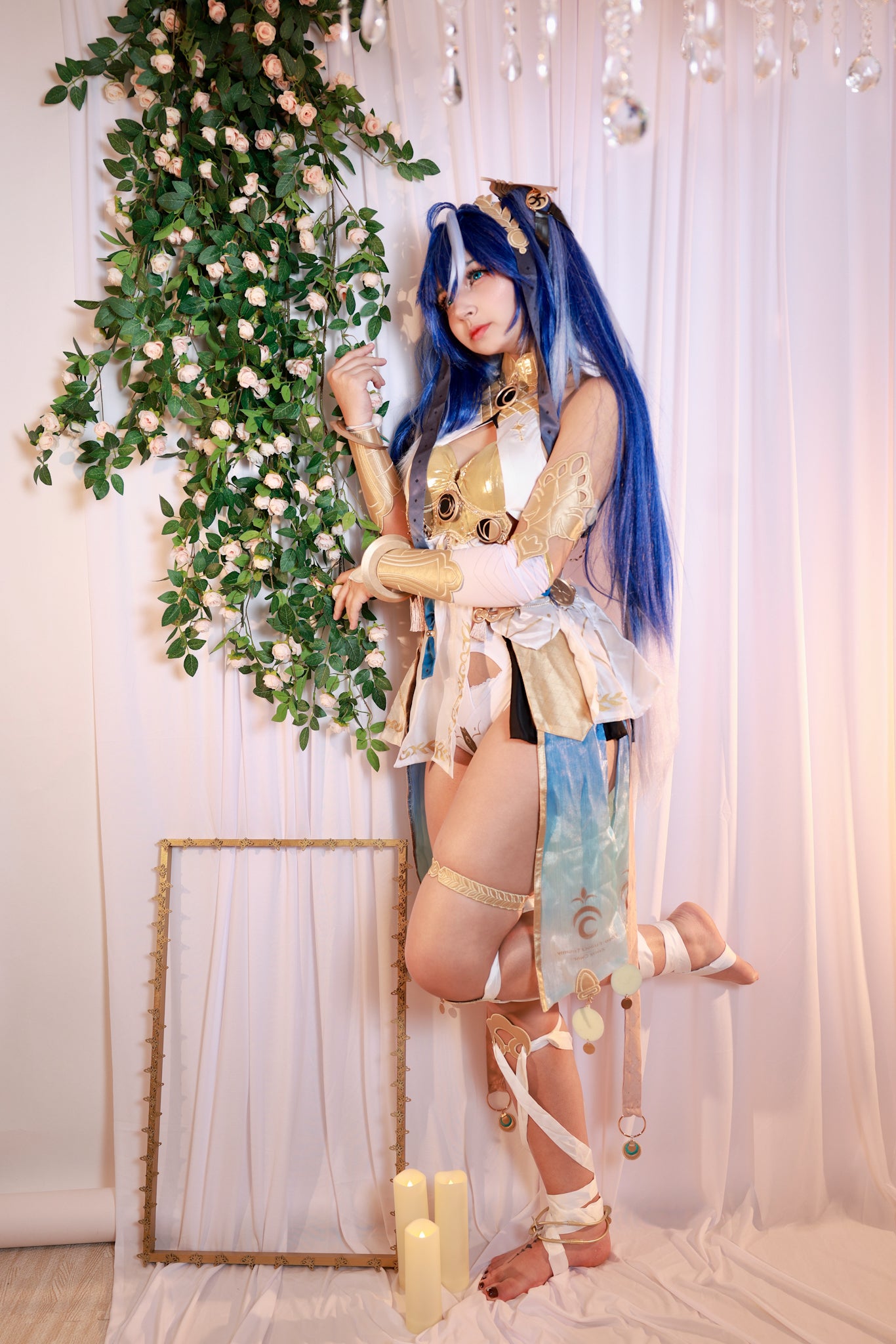 Person in elaborate costume with blue hair standing next to a decorative plant and framed mirror.